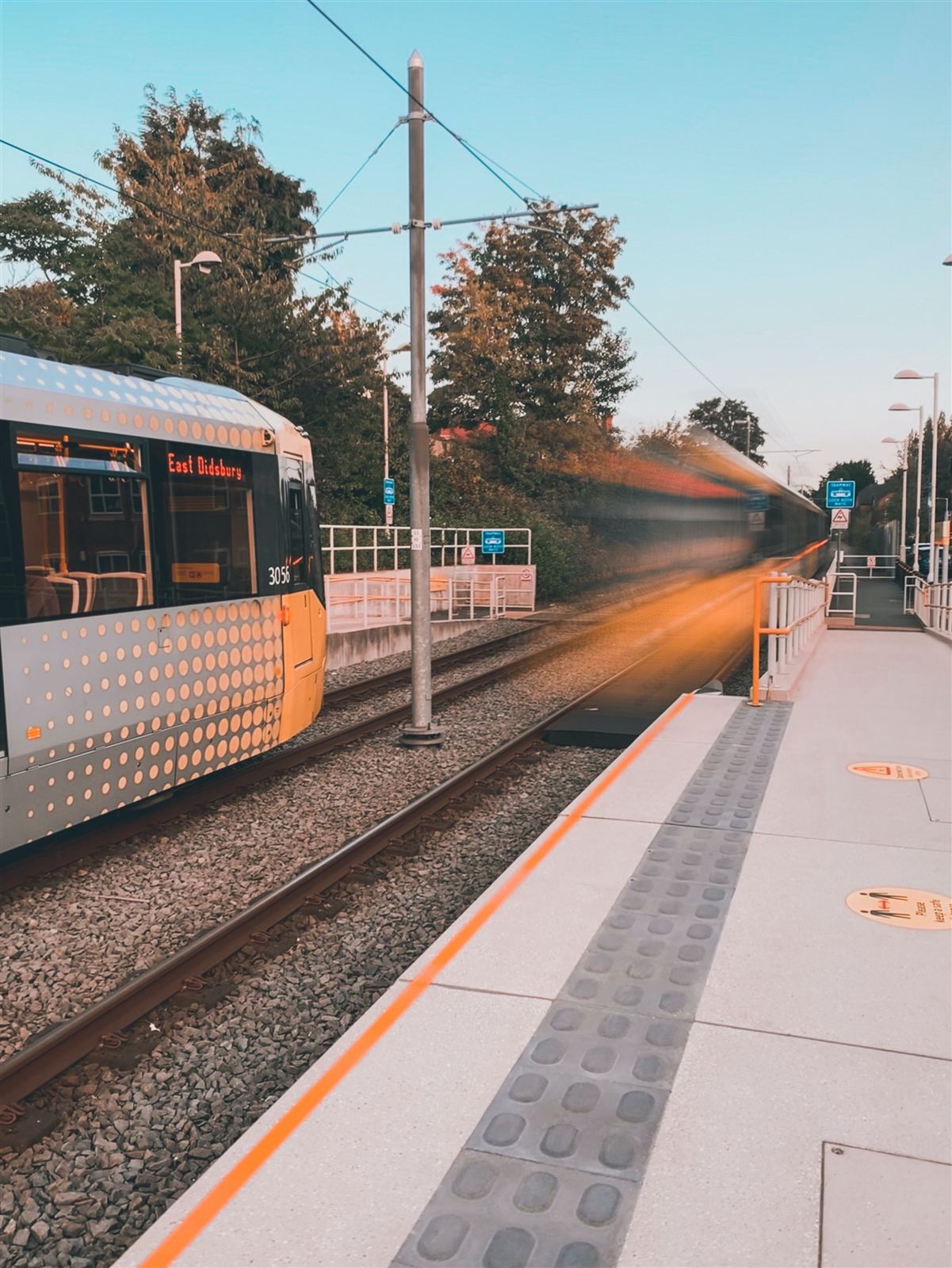 A tram at a suburban stop with a second tram passing in motion blur