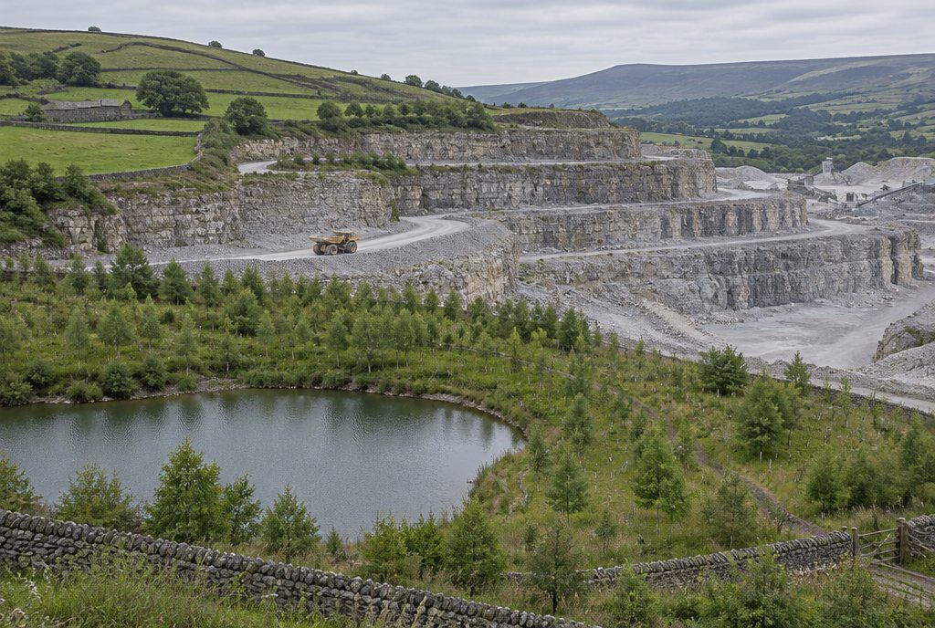 A stepped limestone quarry with a dump truck on the haul road and restored woodland and a lake in the foreground