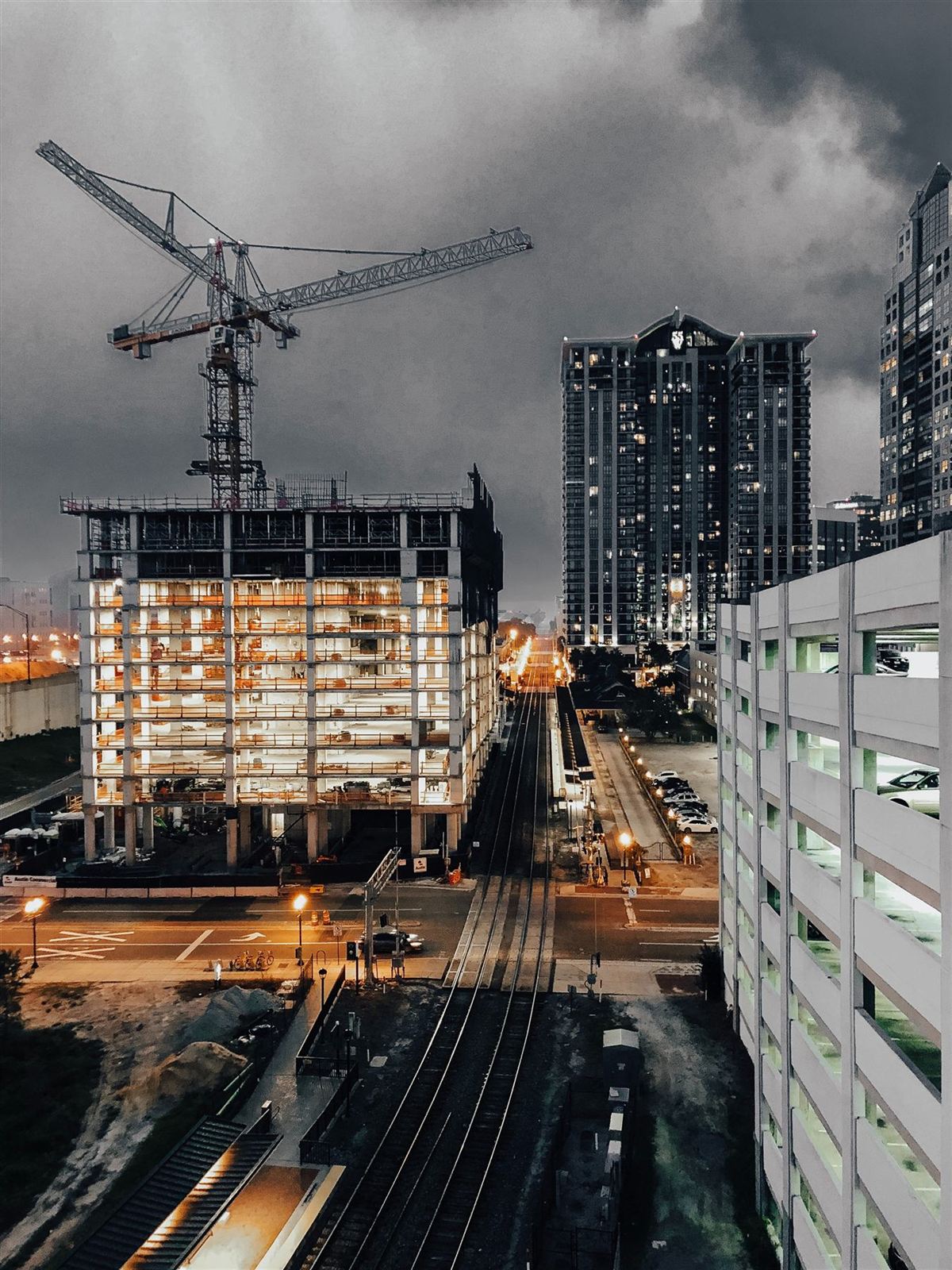 A tower crane over a lit mid-rise building under construction beside a railway at dusk